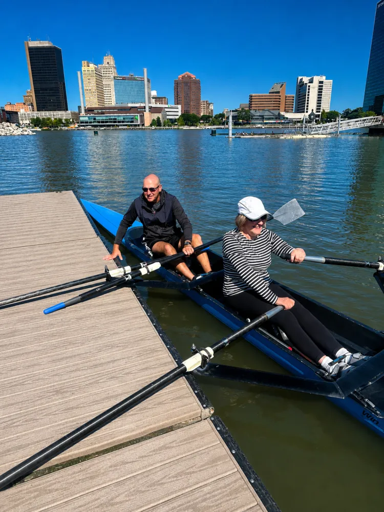 A coach and a rower in a blue double scull at the dock on the Maumee River with the downtown Toledo skyline behind