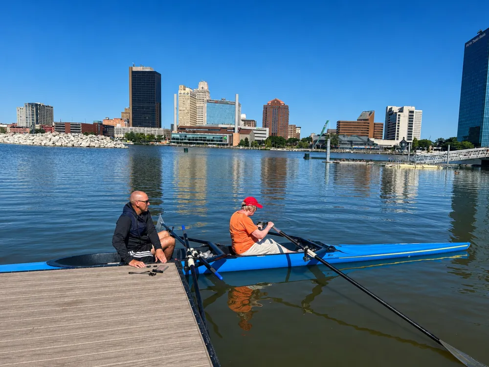 A coach coaching a rower seated in a blue double scull beside the dock on the Maumee River in downtown Toledo